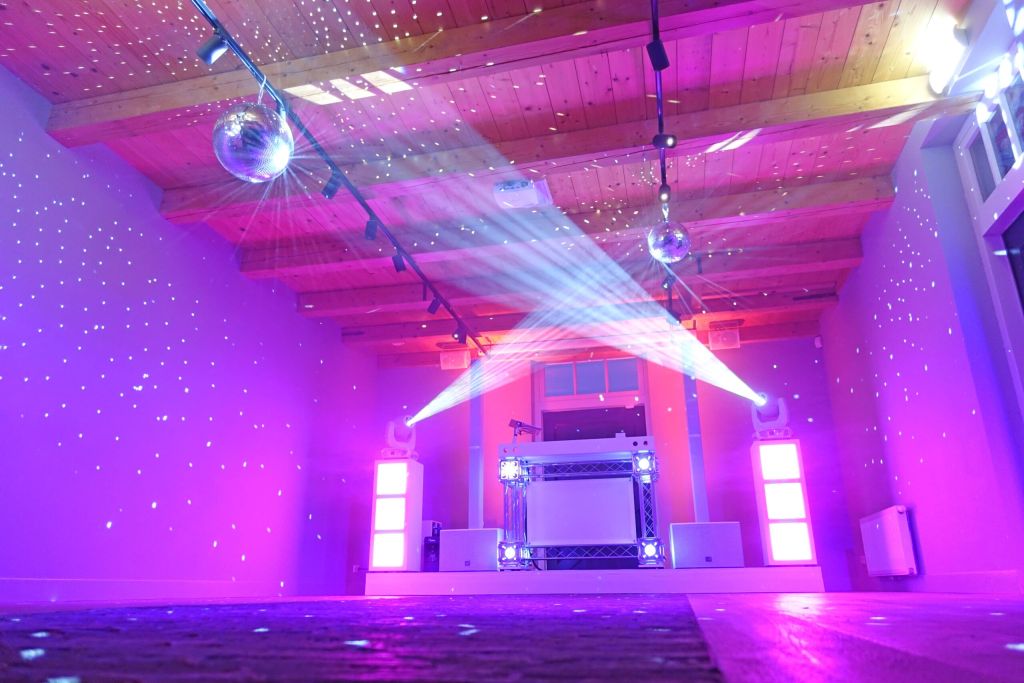Lege ruimte met houten plafond, twee discoballen en DJ-booth, verlicht met paarse en witte lichten.