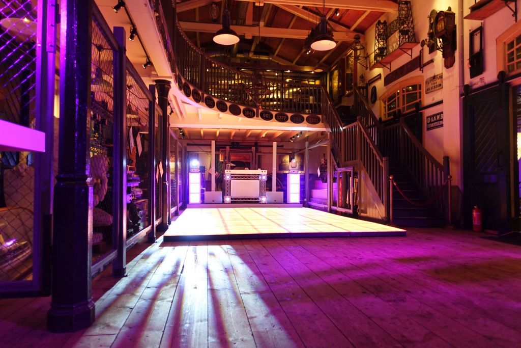 Zaal met houten vloer, verlichte dansvloer, DJ-booth, bovenverdieping en paars-gele sfeerverlichting.